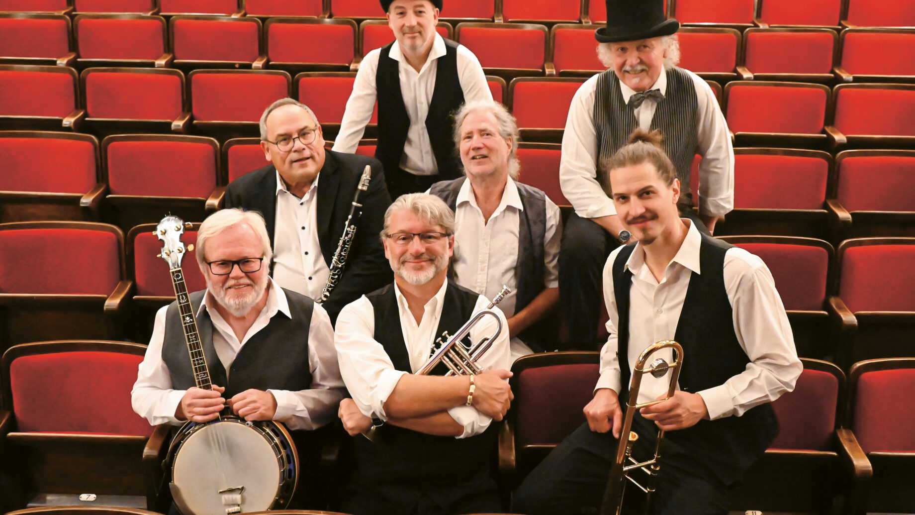 Sieben Männer in Westen und Hemden posieren mit Musikinstrumenten in einem leeren Theater mit roten Sitzen. Einige halten ein Banjo, eine Trompete und eine Posaune.