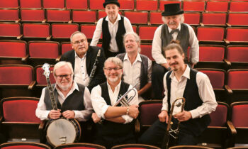 Sieben Männer in Westen und Hemden posieren mit Musikinstrumenten in einem leeren Theater mit roten Sitzen. Einige halten ein Banjo, eine Trompete und eine Posaune.