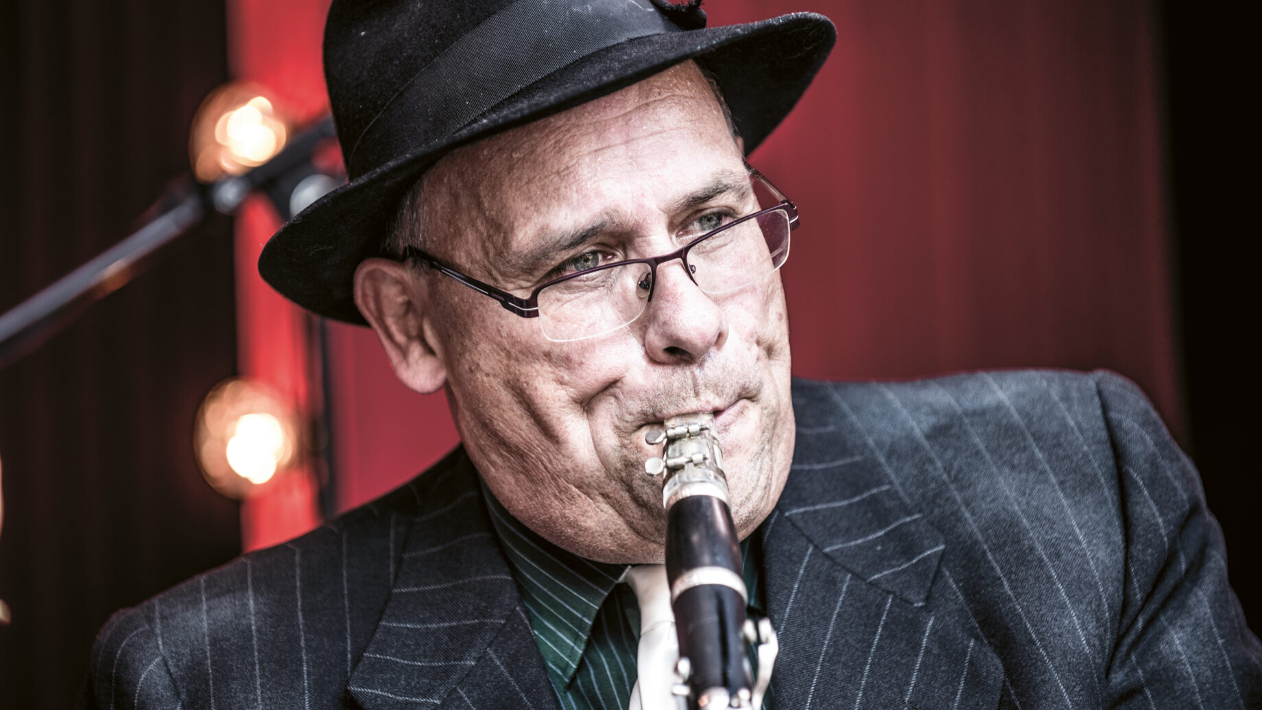 Mann im Nadelstreifenanzug, mit Brille und Filzhut, der Klarinette spielt, vor einem unscharfen Lichterhintergrund.