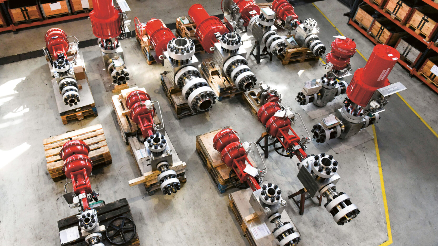Industriearmaturen und Stellantriebe, vor allem in den Farben Rot und Silber, sind auf Holzpaletten in einem Lagerhaus angeordnet, in dessen Nähe sich Regale und Lagerkästen befinden.