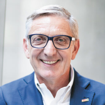 Lächelnder älterer Mann mit grauem Haar und Brille, der einen blauen Anzug und ein weißes Hemd trägt, steht vor einem schlichten hellen Hintergrund.