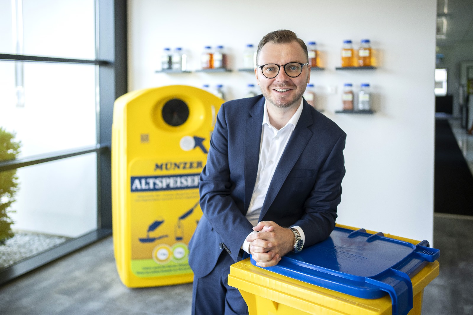 Ein lächelnder Mann im Anzug steht in einer Halle und hebt die bundesländerübergreifende Kooperation im Gesundheitswesen durch eine gelb-blaue Recycling-Tonne hervor.