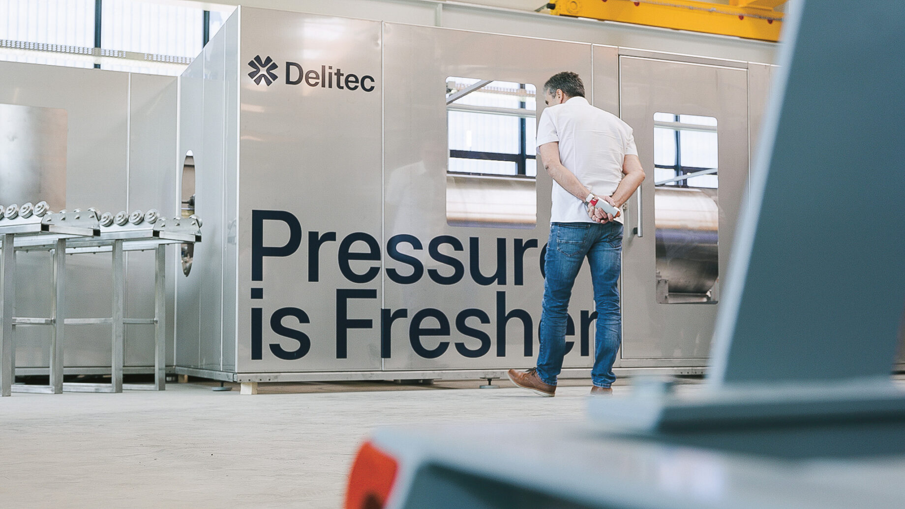 Ein Mann in Jeans steht vor einer großen Metallmaschine mit der Aufschrift "Delitec" und "Pressure is Fresher" in einer hellen Industrieanlage.