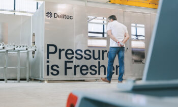 Ein Mann in Jeans steht vor einer großen Metallmaschine mit der Aufschrift "Delitec" und "Pressure is Fresher" in einer hellen Industrieanlage.
