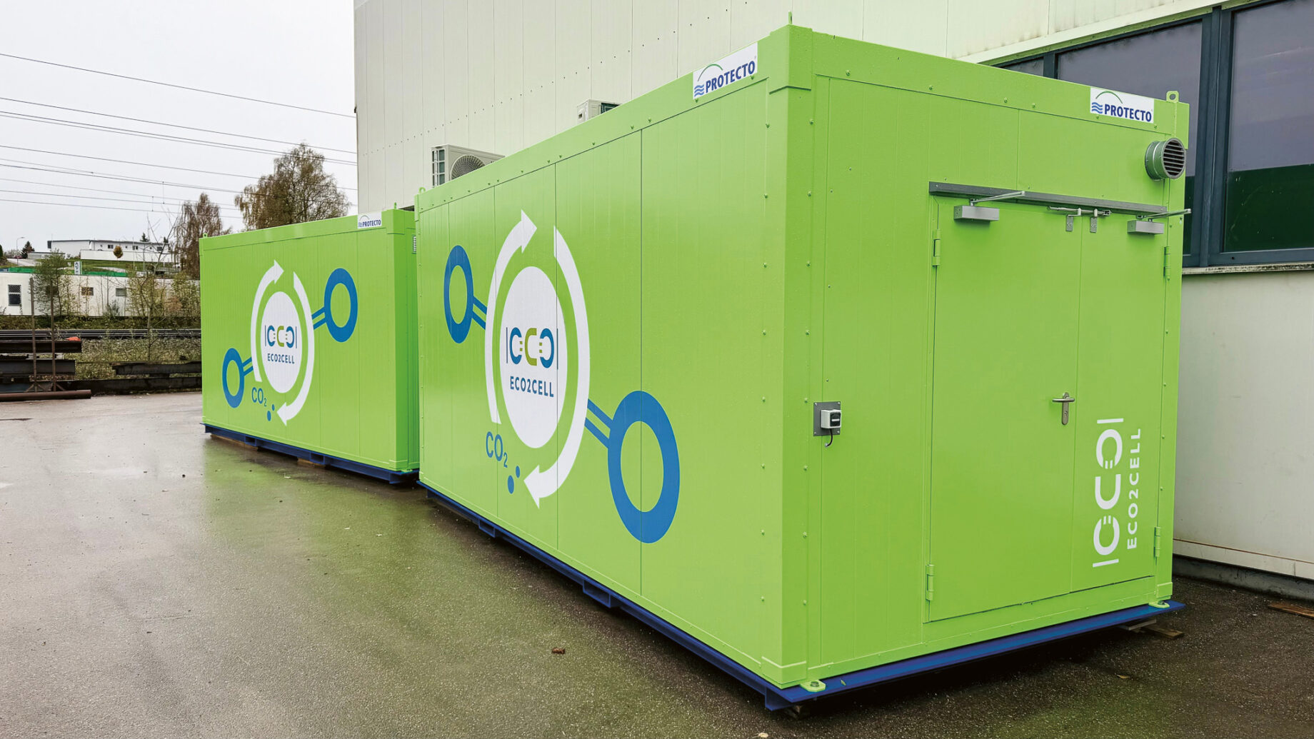 Zwei große grüne Behälter mit der Aufschrift "ECOCELL" und kreisförmigen Grafiken, die im Freien neben einem Gebäude auf einem nassen Bürgersteig aufgestellt sind.