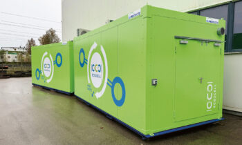 Zwei große grüne Behälter mit der Aufschrift "ECOCELL" und kreisförmigen Grafiken, die im Freien neben einem Gebäude auf einem nassen Bürgersteig aufgestellt sind.