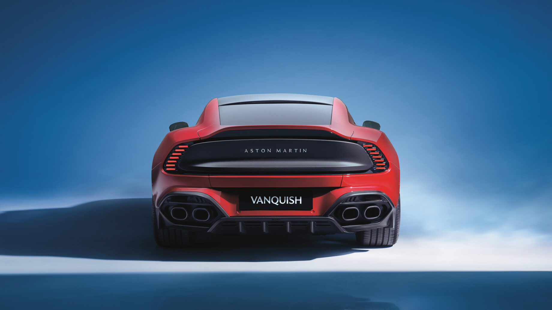Ein roter Aston Martin Vanquish Sportwagen, von hinten gesehen, vor einem blauen Hintergrund mit Farbverlauf, der das schnittige Design und die Rücklichter zur Geltung bringt.