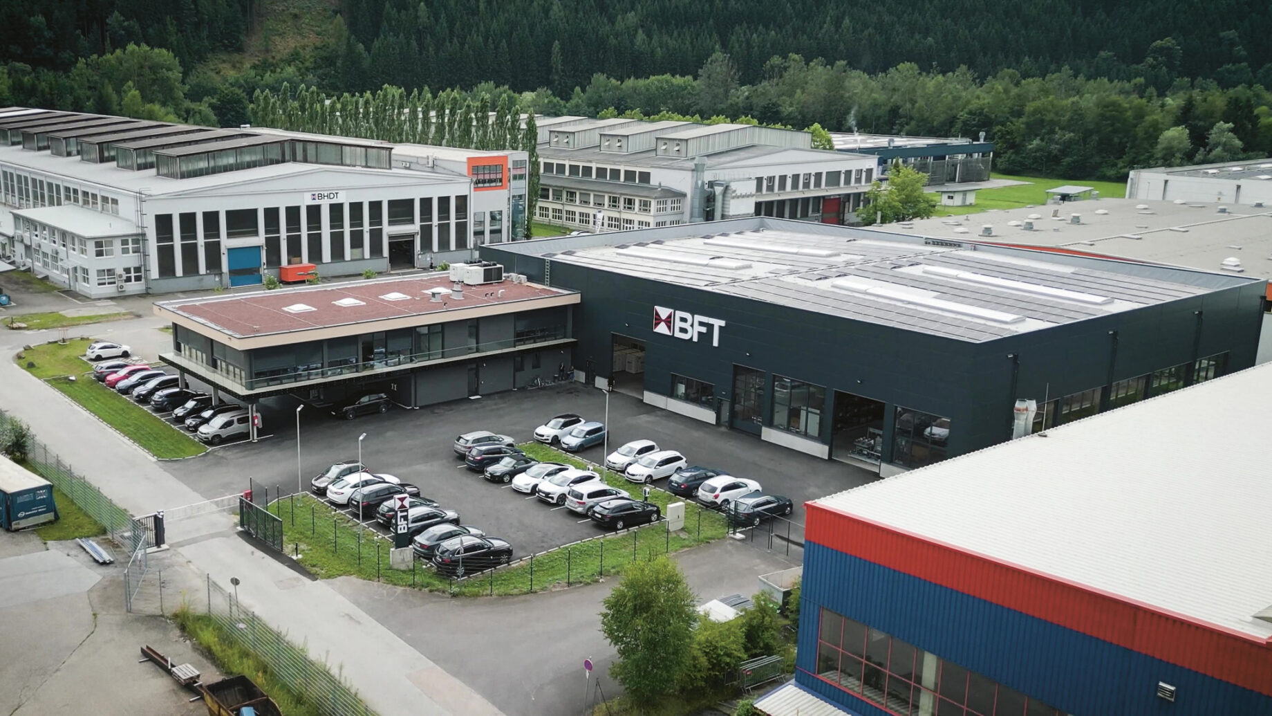 Luftaufnahme eines Industriekomplexes mit mehreren Gebäuden, parkenden Autos, Grünanlagen und einem großen schwarzen Gebäude mit der Aufschrift "BFT" in der Mitte.