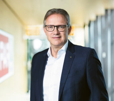 Ein Mann in einem dunklen Anzug und mit Brille steht lächelnd in einem Haus, mit unscharfem Bürohintergrund und einem rot-weißen Schild an der Wand.