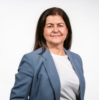 Eine Frau mit langen braunen Haaren trägt einen blauen Blazer und ein weißes Hemd und lächelt vor einem schlichten weißen Hintergrund.