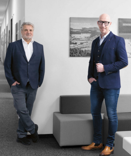 Zwei Männer in Anzügen stehen in einem modernen Büro mit kunstvollen Gemälden hinter ihnen.