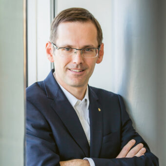 Ein Mann in einem marineblauen Anzug und mit Brille steht mit verschränkten Armen an einem Fenster und lächelt selbstbewusst.