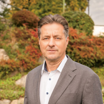 Ein Mann in einem grauen Anzug steht im Freien vor einem Garten mit grünen Sträuchern und Steinen und schaut mit neutralem Blick in die Kamera.