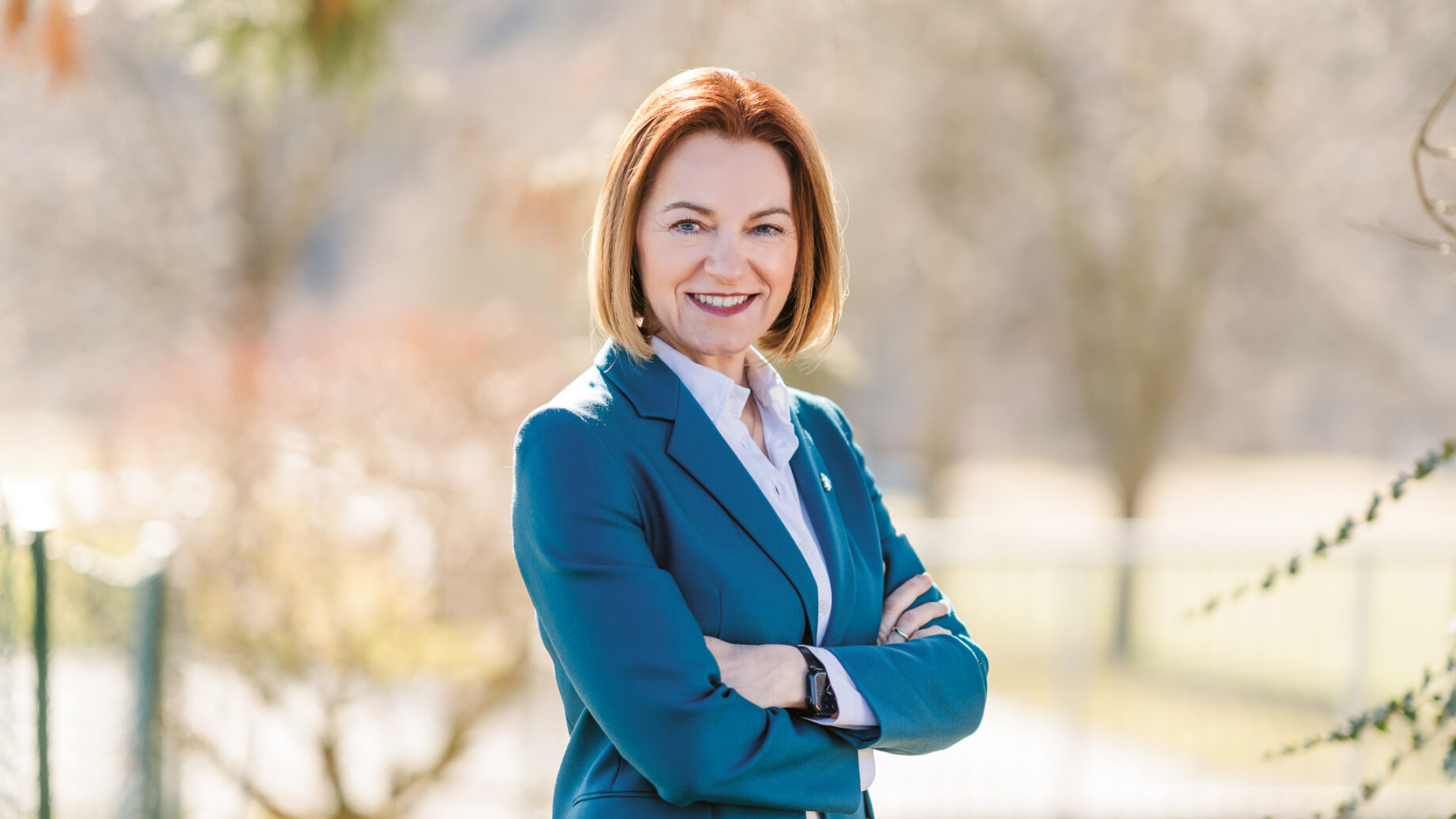 Eine Frau in einem blaugrünen Blazer steht im Freien mit verschränkten Armen und lächelt selbstbewusst in die Kamera. Der natürliche Hintergrund ist unscharf.