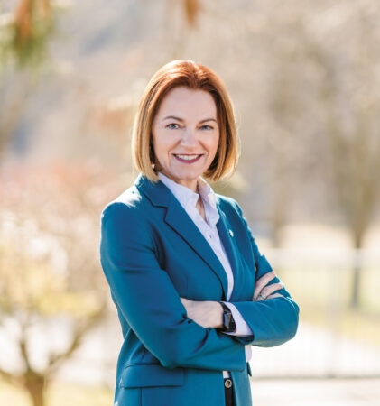 Eine Frau in einem blaugrünen Blazer steht im Freien mit verschränkten Armen und lächelt selbstbewusst in die Kamera. Der natürliche Hintergrund ist unscharf.