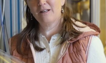 Frau mit langen braunen Haaren in einem Labor. Sie trägt ein weißes Hemd, eine rosa Weste und blaue Handschuhe und hält eine durchsichtige Plastikfolie.