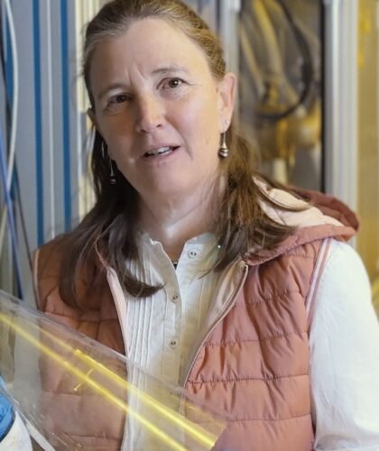 Frau mit langen braunen Haaren in einem Labor. Sie trägt ein weißes Hemd, eine rosa Weste und blaue Handschuhe und hält eine durchsichtige Plastikfolie.