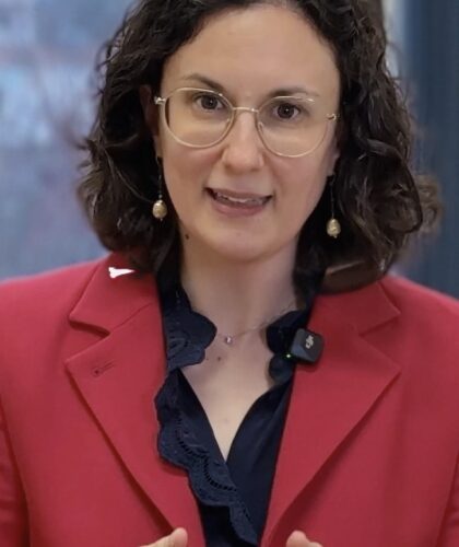 Eine Frau mit braunen haaren und roter Weste steht in einem Raum und spricht.