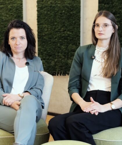 Zwei Frauen, sitzen in Geschäftskleidung nebeneinander in einem modernen Büro mit grünen Wandpaneelen hinter ihnen.