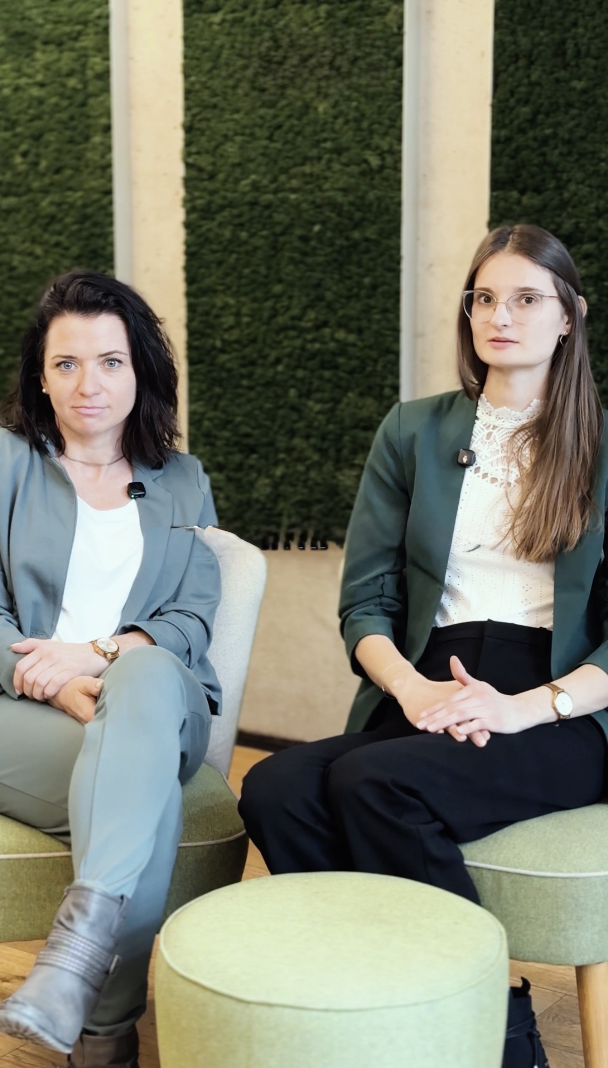Zwei Frauen, sitzen in Geschäftskleidung nebeneinander in einem modernen Büro mit grünen Wandpaneelen hinter ihnen.