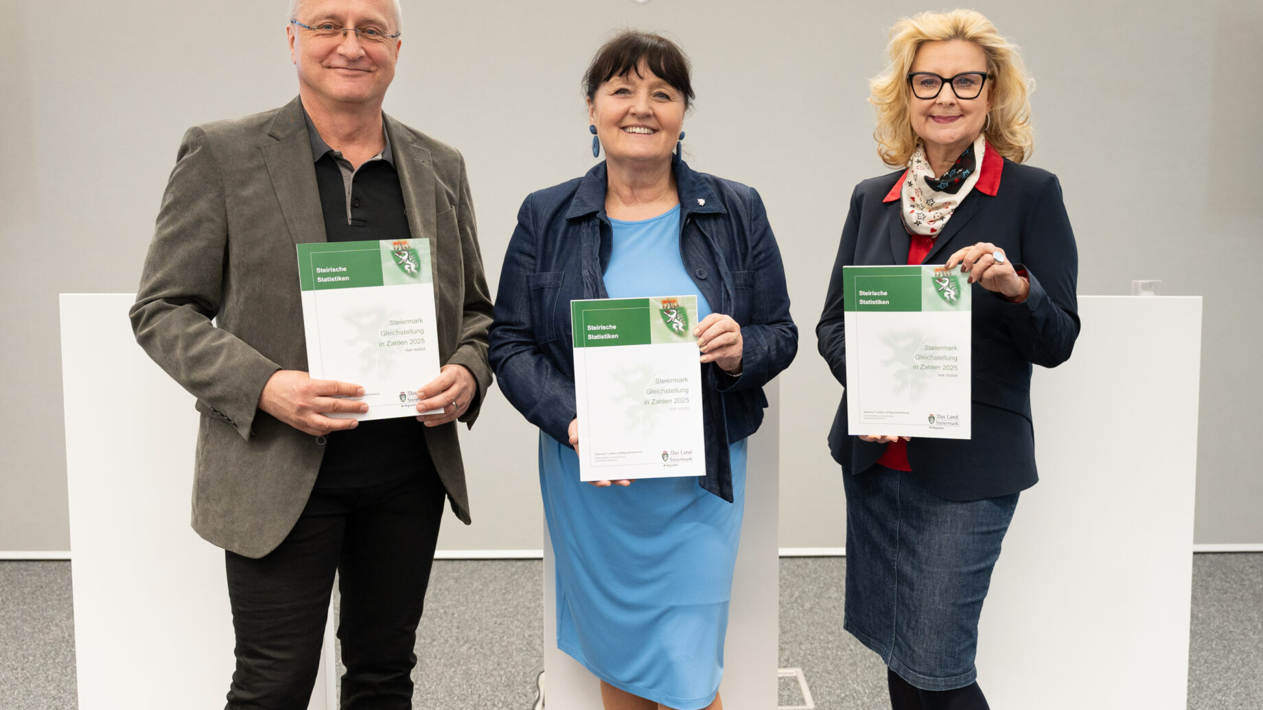 Drei lächelnde Erwachsene mit Urkunden in Business-Kleidung stehen in der Halle - als Symbol für die klare Mehrheit für soziale Sicherheit in der Steiermark.