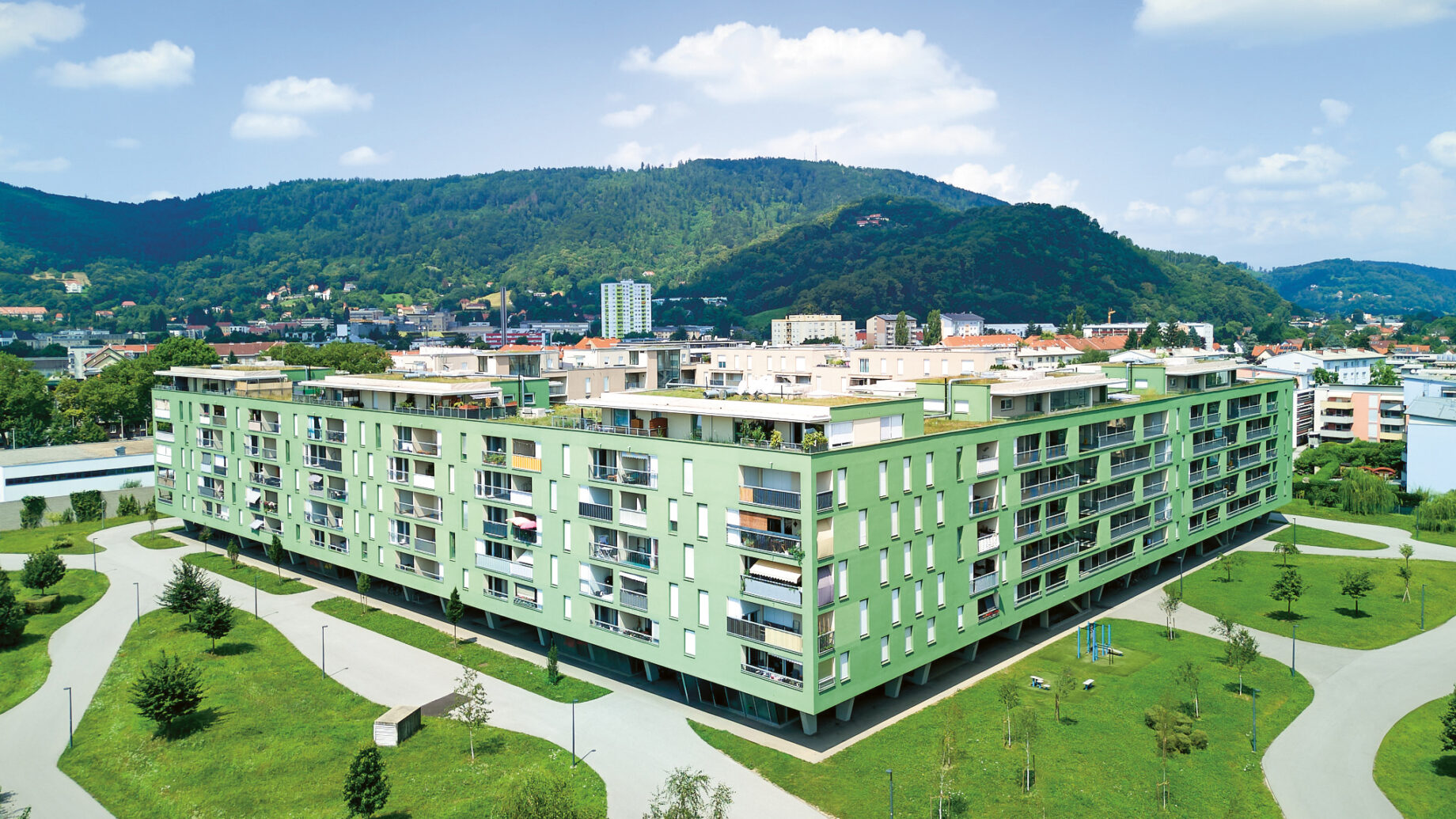 Ein großes, modernes, grünes Apartmenthaus mit Balkonen, umgeben von Wegen, Gras und Bäumen, vor einer Kulisse aus Hügeln und blauem Himmel.