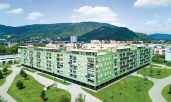 Ein großes, modernes, grünes Apartmenthaus mit Balkonen, umgeben von Wegen, Gras und Bäumen, vor einer Kulisse aus Hügeln und blauem Himmel.