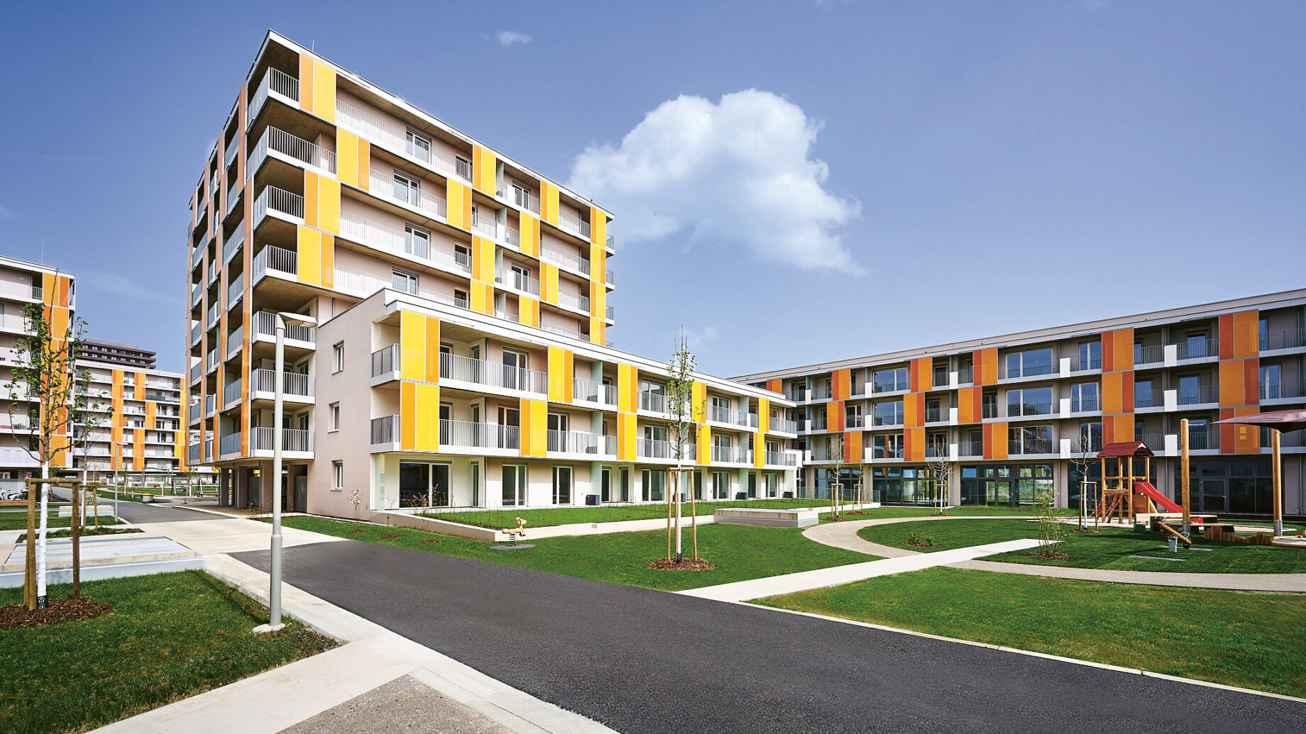 Moderne Wohngebäude mit gelben und orangefarbenen Akzenten, umgeben von grünen Rasenflächen, Gehwegen und einem kleinen Spielplatz unter blauem Himmel.