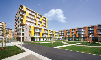 Moderne Wohngebäude mit gelben und orangefarbenen Akzenten, umgeben von grünen Rasenflächen, Gehwegen und einem kleinen Spielplatz unter blauem Himmel.