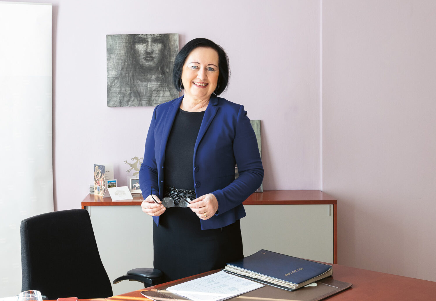 Eine Frau in Geschäftskleidung steht lächelnd in einem Büro mit einem Schreibtisch, Dokumenten und einem Transparent mit der Aufschrift "Bildungsdirektion Steiermark" im Hintergrund.