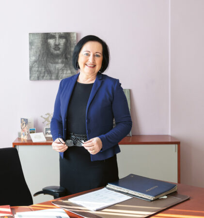 Eine Frau in Geschäftskleidung steht lächelnd in einem Büro mit einem Schreibtisch, Dokumenten und einem Transparent mit der Aufschrift "Bildungsdirektion Steiermark" im Hintergrund.