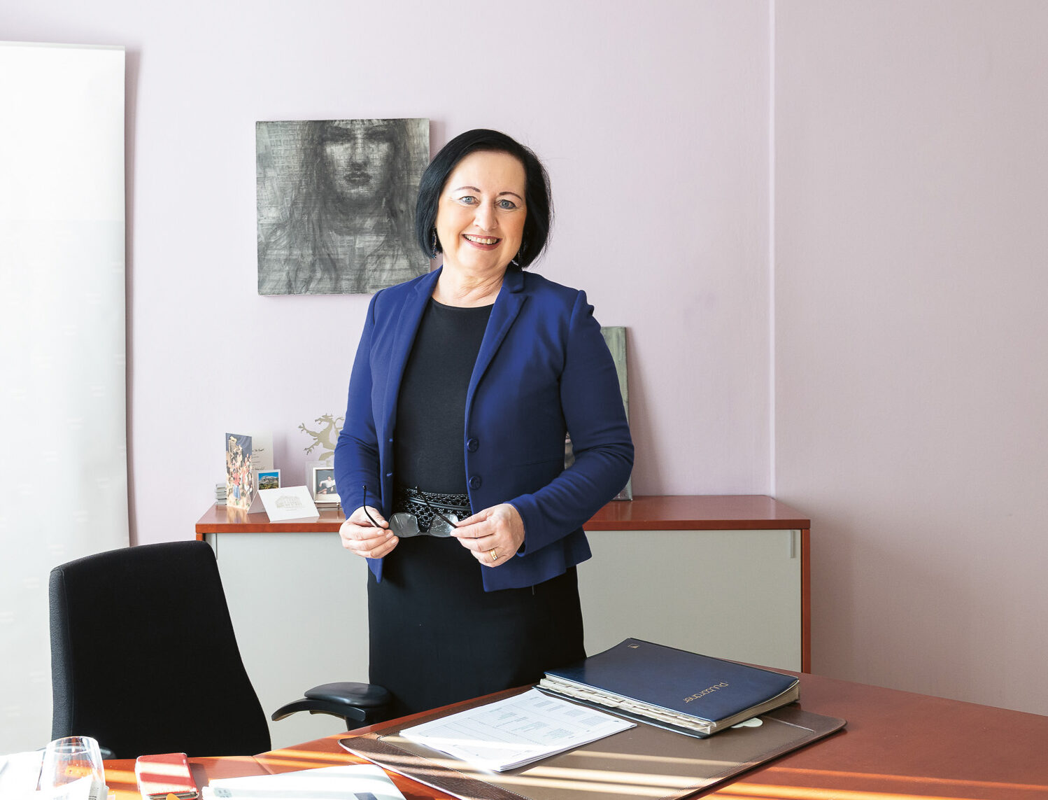 Eine Frau in Geschäftskleidung steht lächelnd in einem Büro mit einem Schreibtisch, Dokumenten und einem Transparent mit der Aufschrift "Bildungsdirektion Steiermark" im Hintergrund.