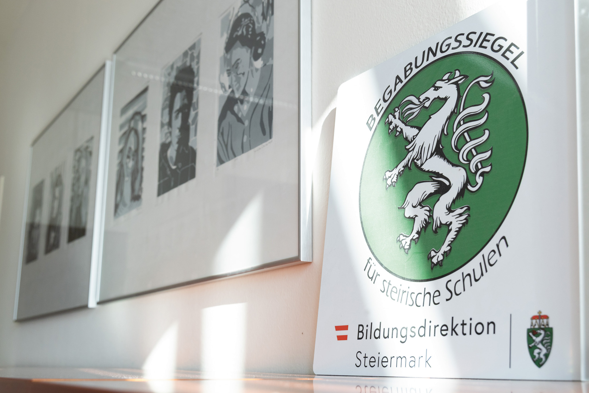 Auf einem Schild mit einem grünen Löwenemblem steht "Begabungssiegel für steirische Schulen" neben gerahmter Schwarz-Weiß-Kunst an einer sonnenbeschienenen Wand.