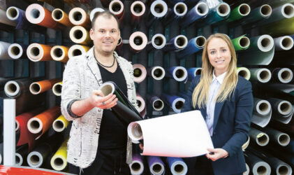 Zwei Personen stehen in einer Werkstatt oder einem Geschäft vor bunten Vinylrollen, halten Stoffbahnen in der Hand und lächeln in die Kamera.