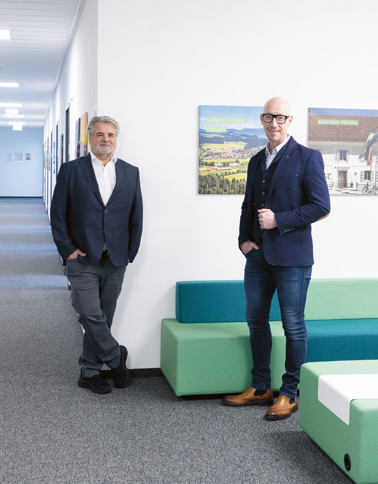 Zwei Männer in Geschäftskleidung stehen in einem modernen Büroflur mit bunten Sitzgelegenheiten und Landschaftsfotos an der Wand.