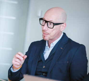 Ein glatzköpfiger Mann mit Brille und blauem Anzug gestikuliert, während er in einer modernen Büroumgebung spricht, in der im Vordergrund Laptops zu sehen sind.