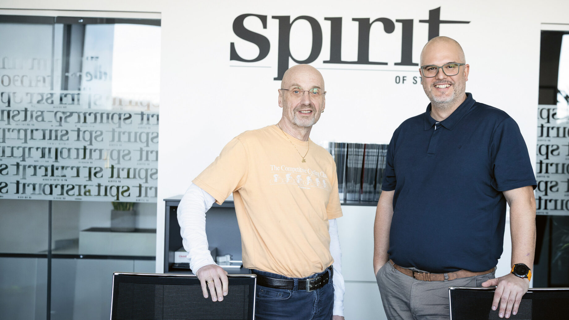 Zwei lächelnde Männer stehen in einem Büro vor einer Wand mit der Aufschrift "Geist", beide tragen Brillen und Freizeitkleidung.