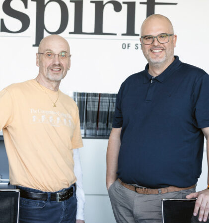 Zwei lächelnde Männer stehen in einem Büro vor einer Wand mit der Aufschrift "Geist", beide tragen Brillen und Freizeitkleidung.