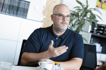 Ein Mann mit Brille und marineblauem Hemd gestikuliert, während er mit einer Tasse Kaffee an einem Tisch in einem Büro sitzt.