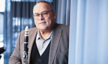 Ein Mann mit Brille und Anzug steht in einem Innenraum und hält eine Klarinette in der Hand. Im Hintergrund sind blaue Vorhänge und sanftes Tageslicht zu sehen.