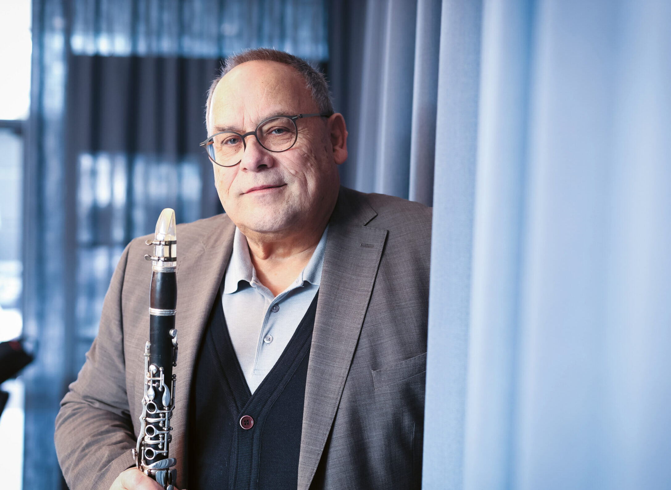 Ein Mann mit Brille und Anzug steht in einem Innenraum und hält eine Klarinette in der Hand. Im Hintergrund sind blaue Vorhänge und sanftes Tageslicht zu sehen.