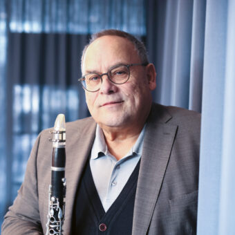 Ein Mann mit Brille und Anzugjacke steht drinnen vor blauen Vorhängen, hält eine Klarinette in der Hand und blickt mit einem leichten Lächeln in die Kamera.