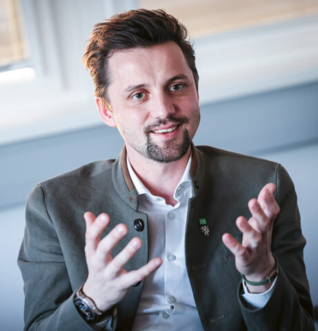 Ein Mann mit kurzen Haaren und einem Bart, der einen Blazer trägt, gestikuliert mit beiden Händen, während er in einem Haus sitzt und lächelt.