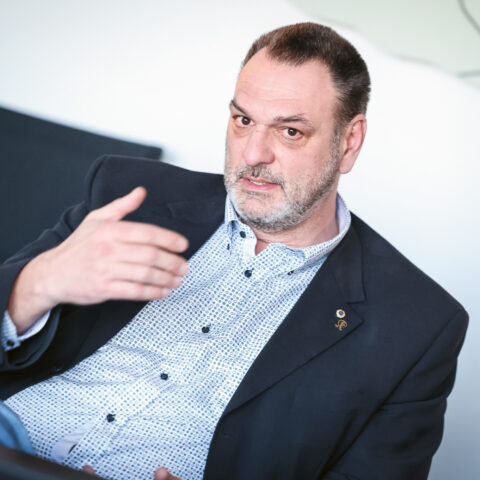Ein Mann in einem Blazer und gemustertem Hemd gestikuliert mit der Hand, während er spricht, und sitzt in einem Haus vor einem hellen Hintergrund.