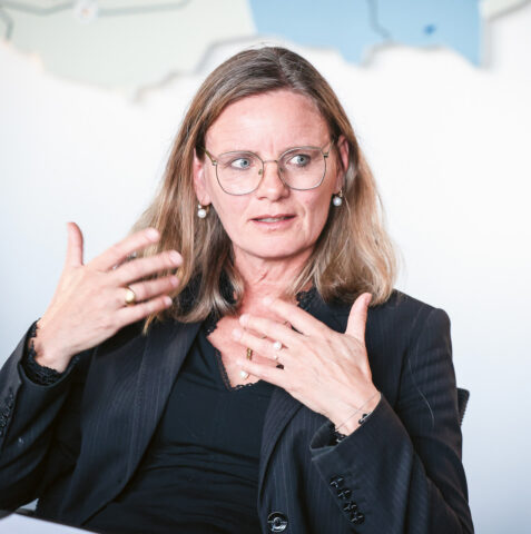 Eine Frau mit Brille und schwarzem Anzug gestikuliert mit den Händen, während sie spricht. Sie sitzt in einem Raum vor einer verschwommenen Landkarte an der Wand.