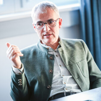 Älterer Mann mit grauem Haar und Brille gestikuliert, während er spricht. Er trägt eine grüne Jacke und ein kariertes Hemd und sitzt in einem Haus an einem Tisch.