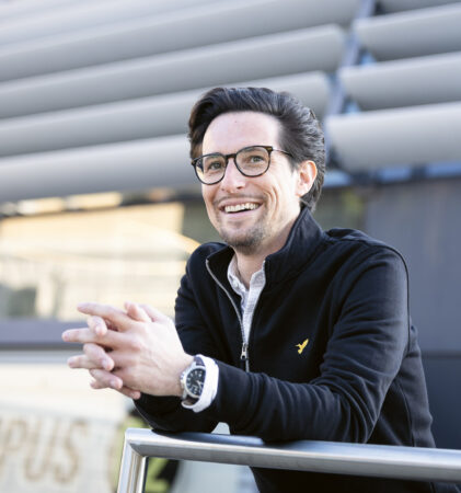 Ein lächelnder Mann mit Brille und schwarzer Jacke lehnt an einem Geländer vor einem modernen Gebäude mit Metallplatten und großen Fenstern.