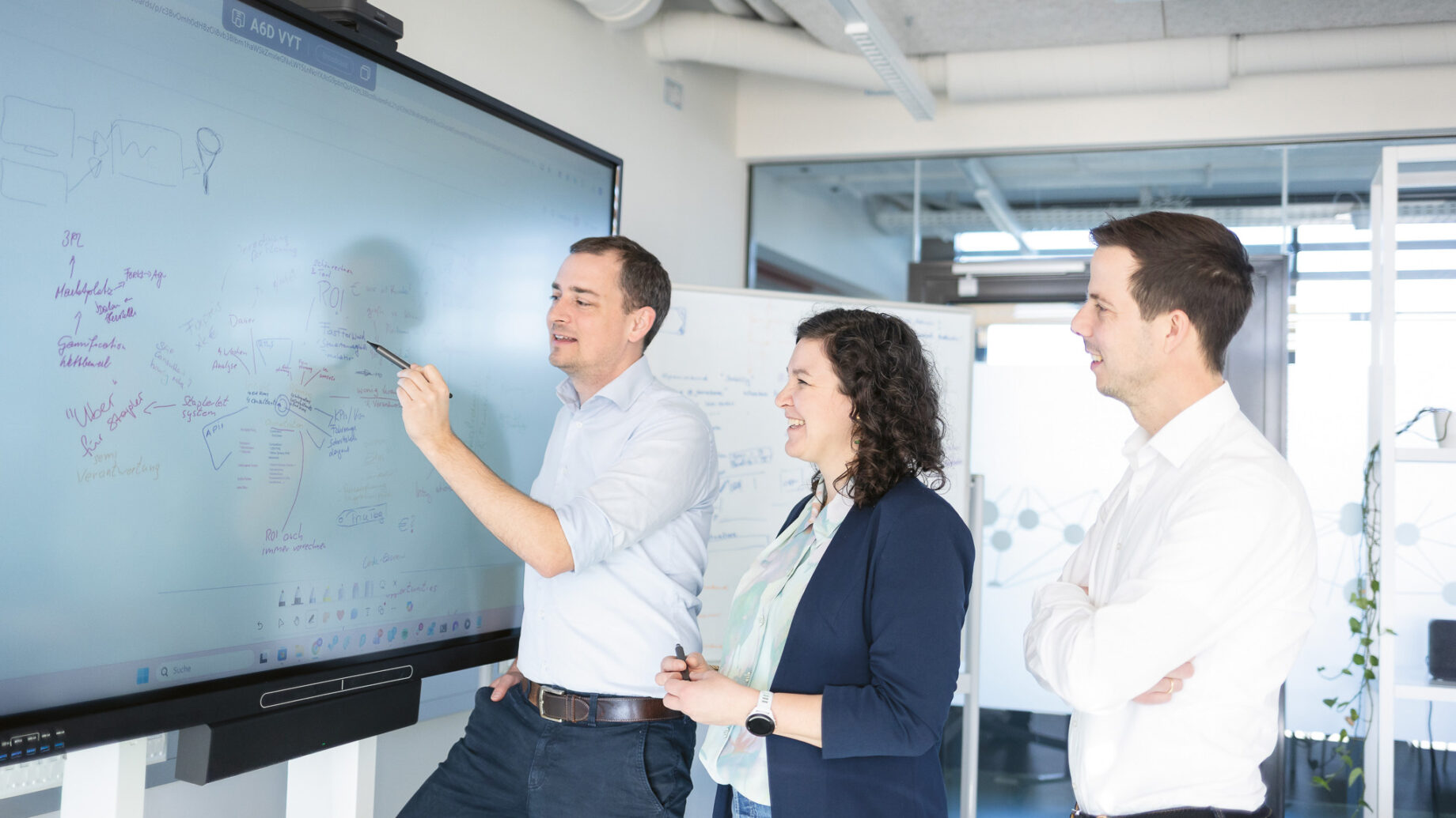 Drei Personen, die an einem großen digitalen Whiteboard stehen und in einem modernen, hellen Büroraum diskutieren und Notizen und Diagramme schreiben.