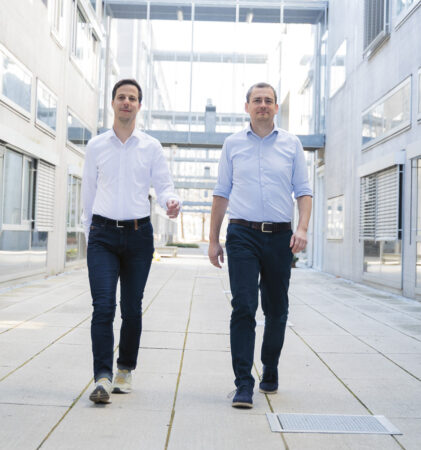 Zwei Männer in weißen Hemden und dunklen Hosen gehen selbstbewusst einen modernen Korridor im Freien entlang, der von Glas- und Betongebäuden umgeben ist.