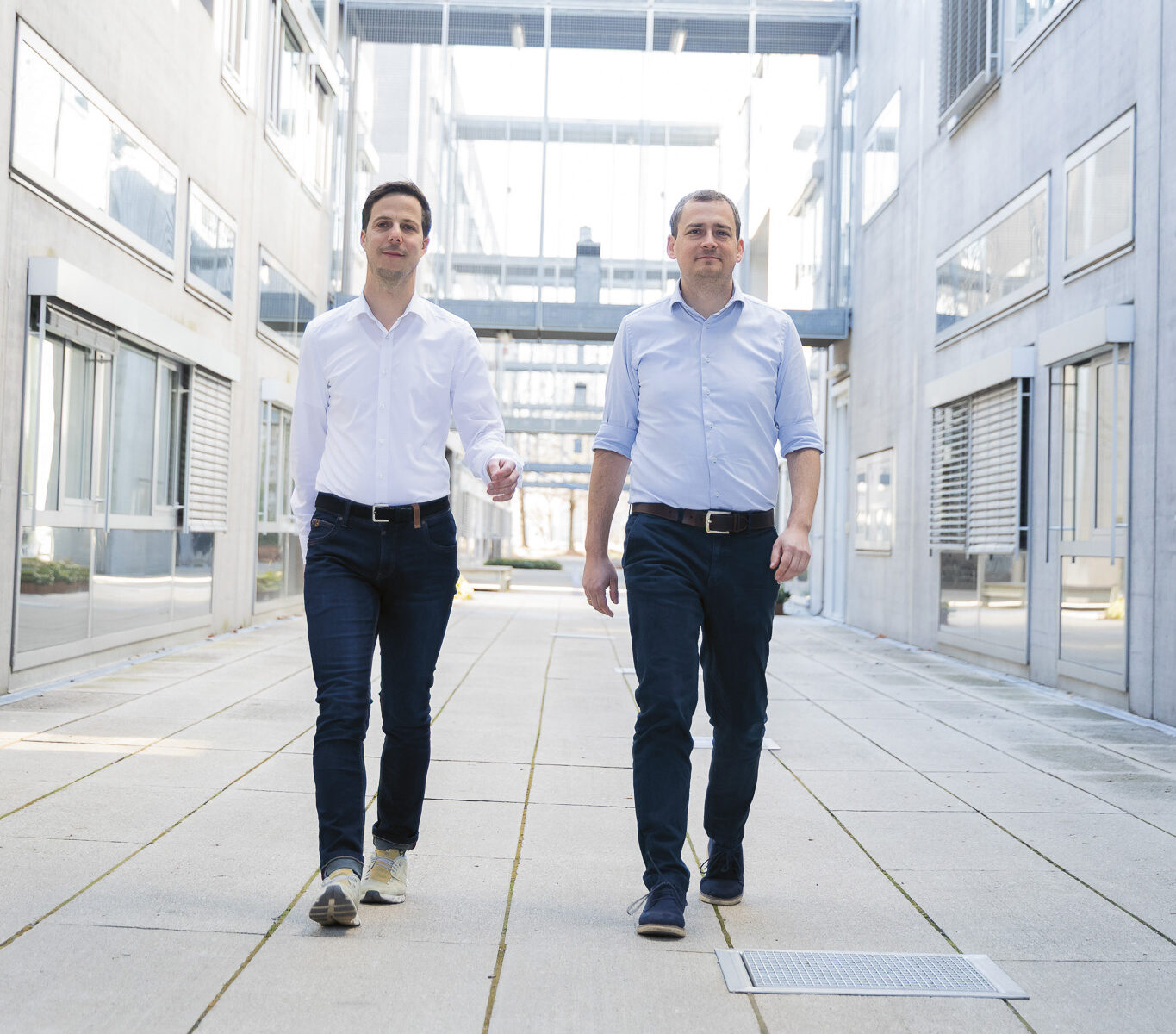 Zwei Männer in weißen Hemden und dunklen Hosen gehen selbstbewusst einen modernen Korridor im Freien entlang, der von Glas- und Betongebäuden umgeben ist.